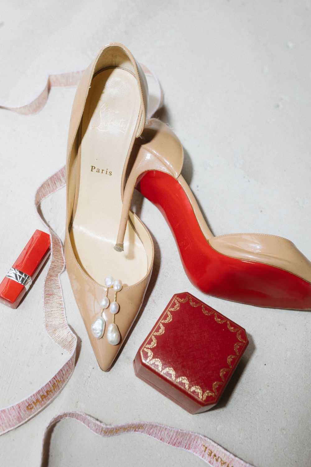 ouboutin shoes, a Cartier ring box, and lipstick displayed for a wedding detail photo, captured with flash.