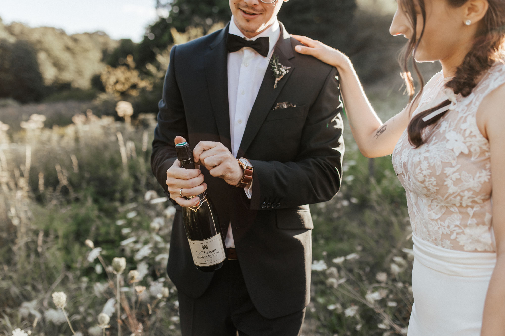 Bride and groom standing together; groom opens champagne bottle.