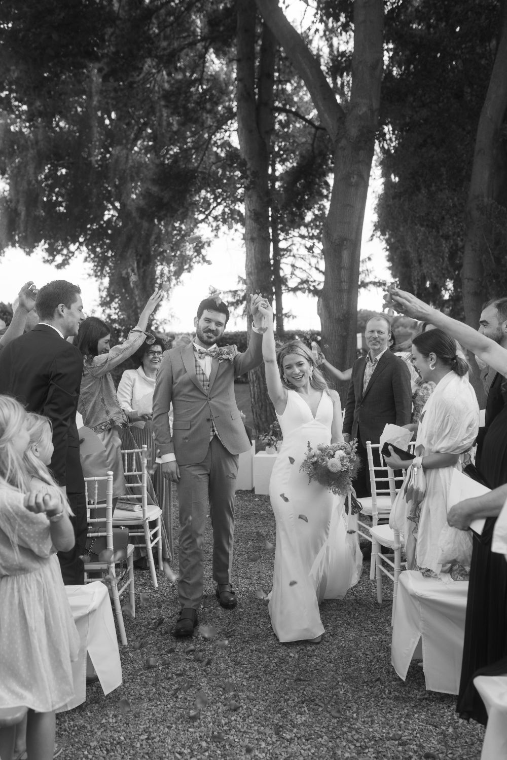 Bride and groom exiting ceremony, hands raised in joy as petals are thrown.