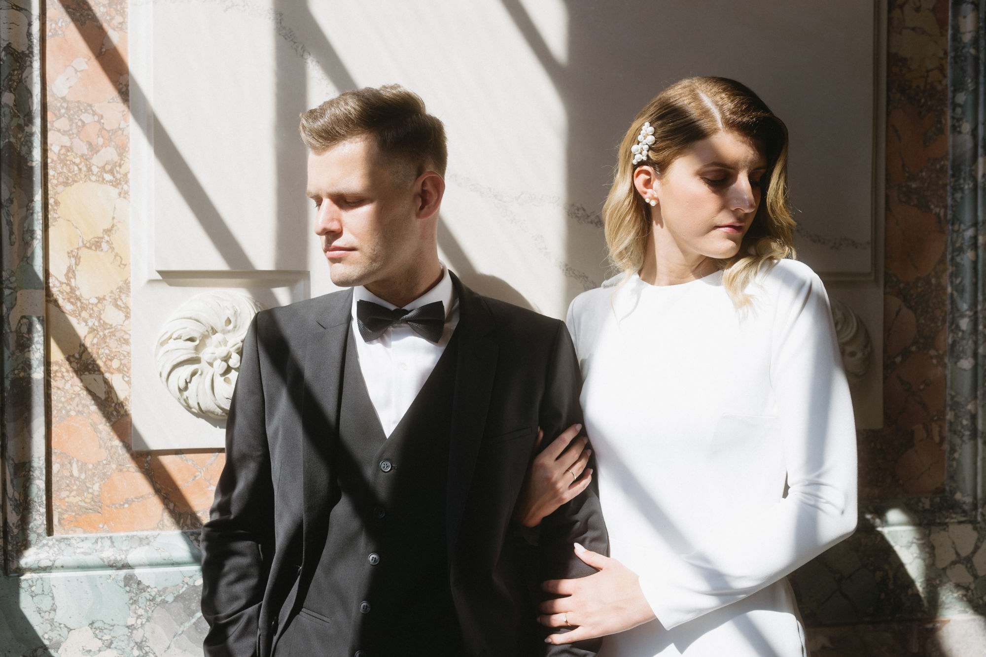 Bride and groom standing together, bathed in light and shadow from a window, looking in different directions.