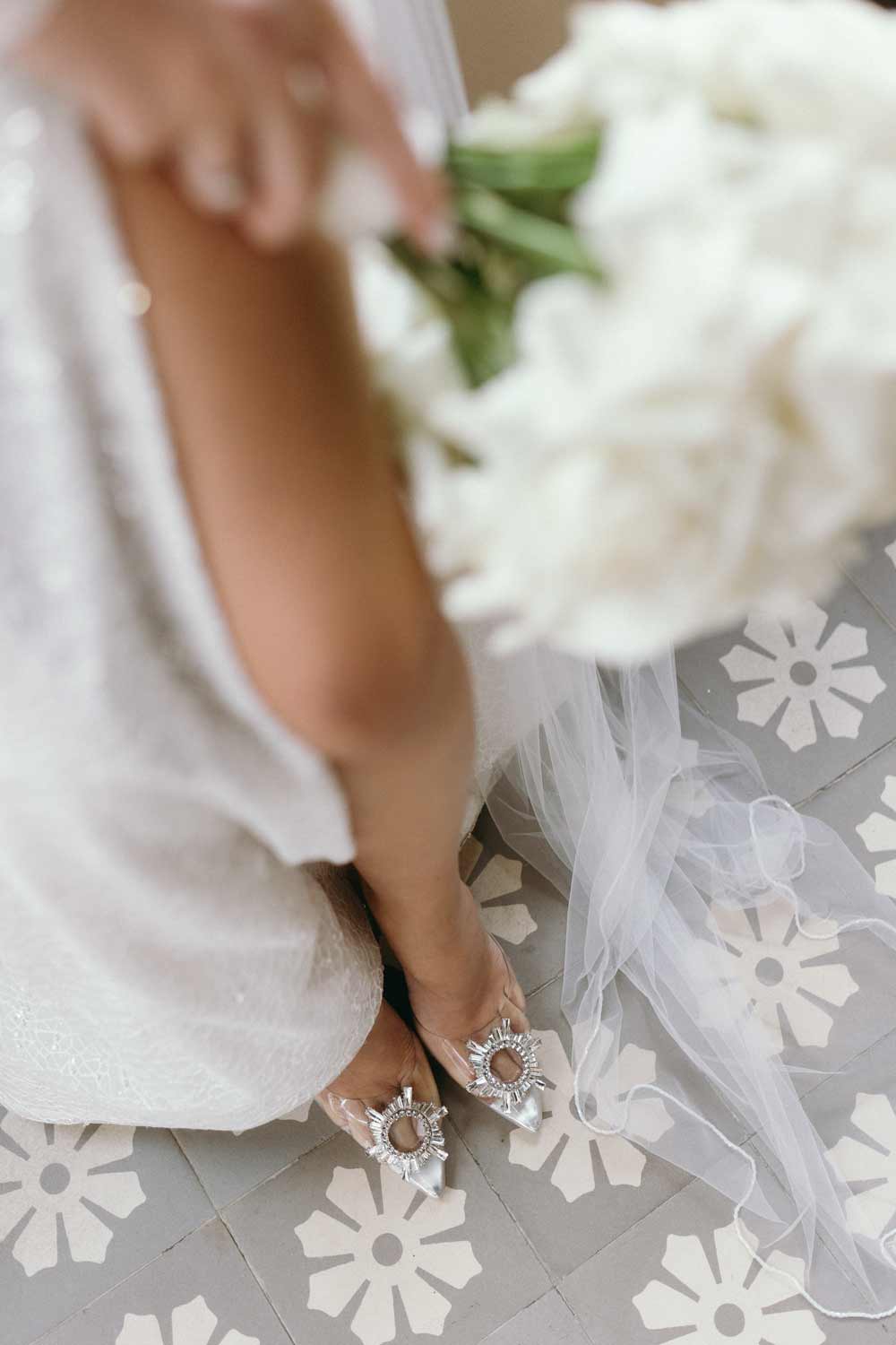 Detail shot of bride's legs in an open-cut dress, holding a bouquet of white roses and wearing Amina Muaddi shoes.