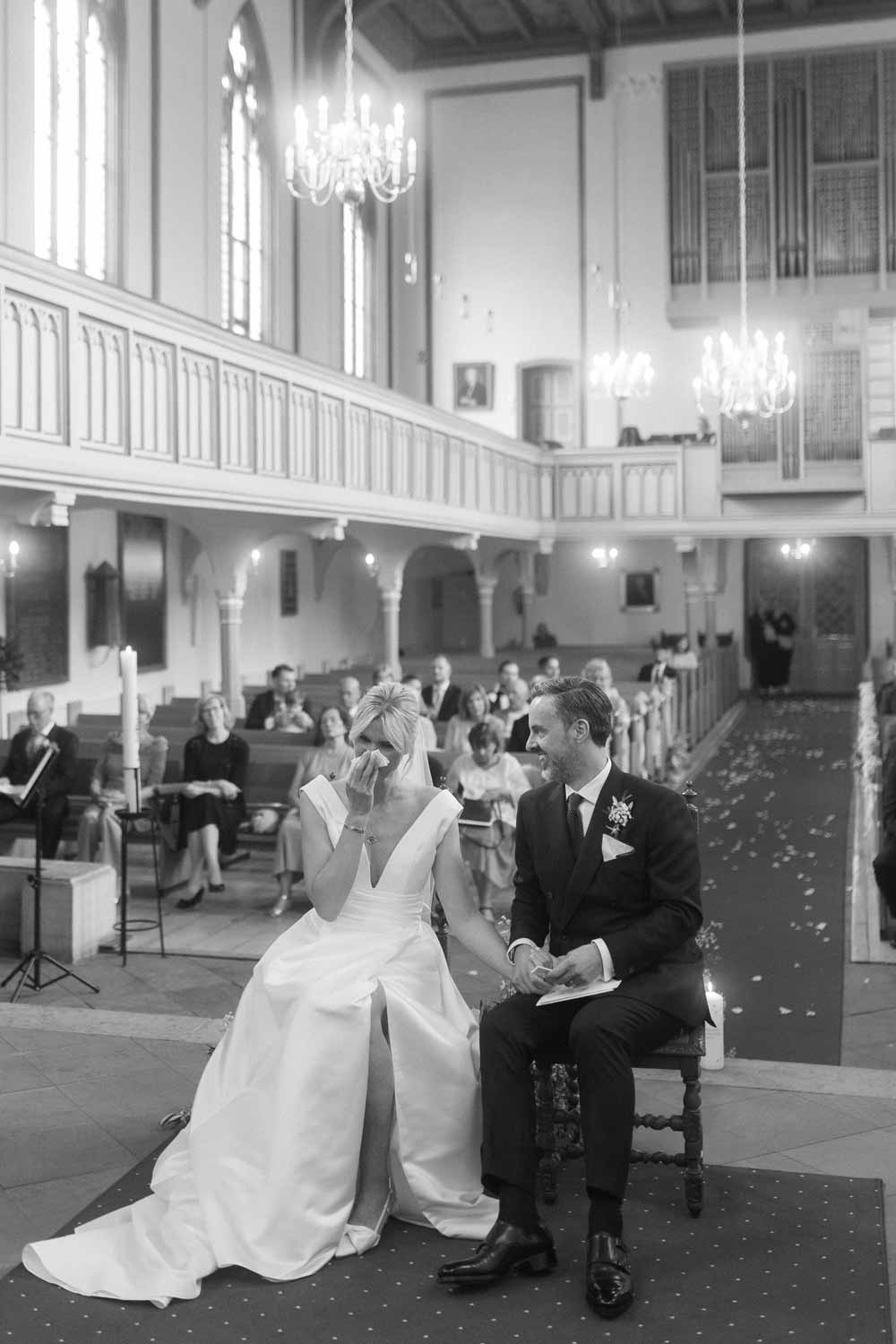 Bride wipes a tear while seated with groom during church ceremony.