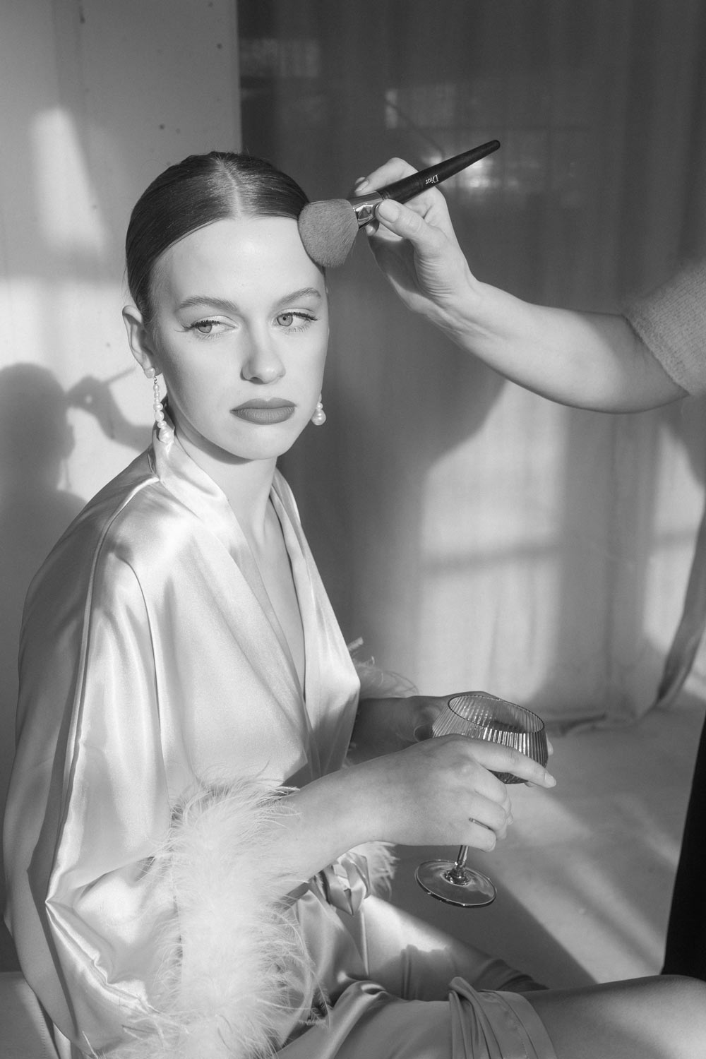 Glamorous bride sits bathed in sunlight, holding champagne, receiving final touches with a Dior brush from her stylist.