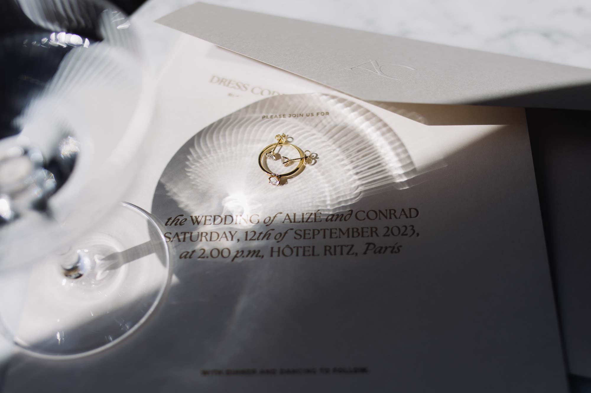 Wedding stationery displayed with soft light through a window, adorned with diamond jewellery and a champagne glass casting shadows.